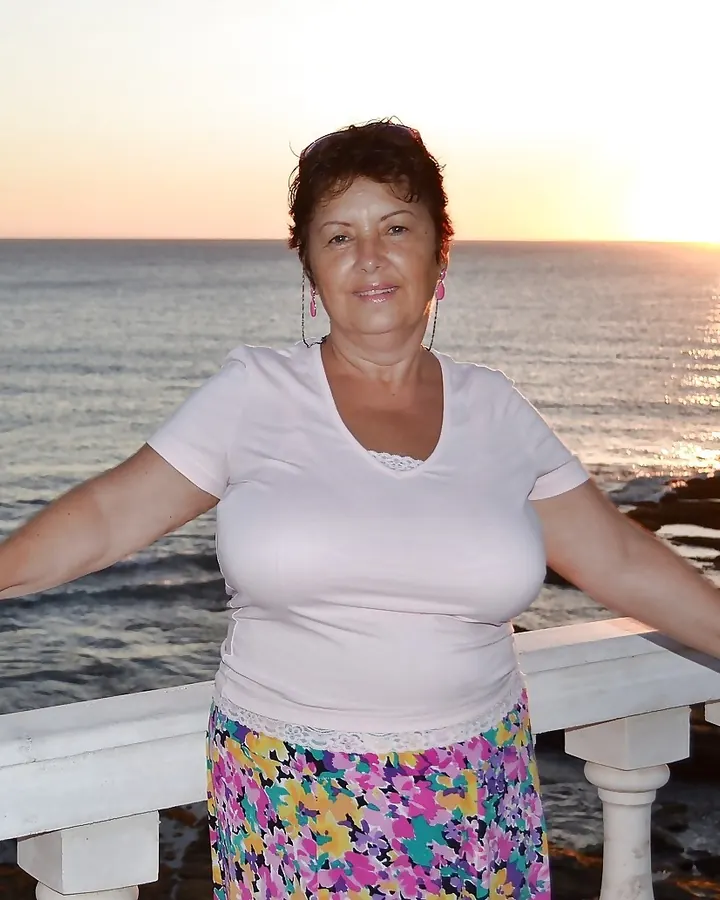 Femme latino en t-shirt blanc et jupe fleurie, debout sur un balcon face à la mer au coucher du soleil