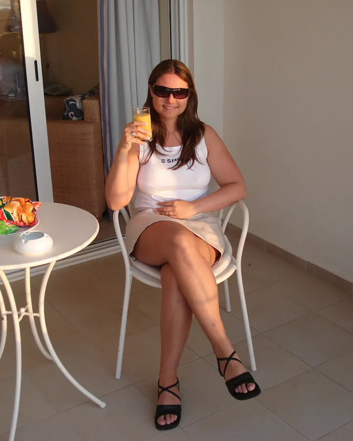 Femme en débardeur blanc et jupe beige, sur une terrasse avec un verre