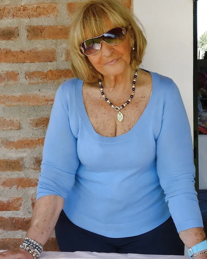 Femme âgée aux cheveux blonds, lunettes de soleil, vêtue d'un pull bleu, assise devant un mur de briques.