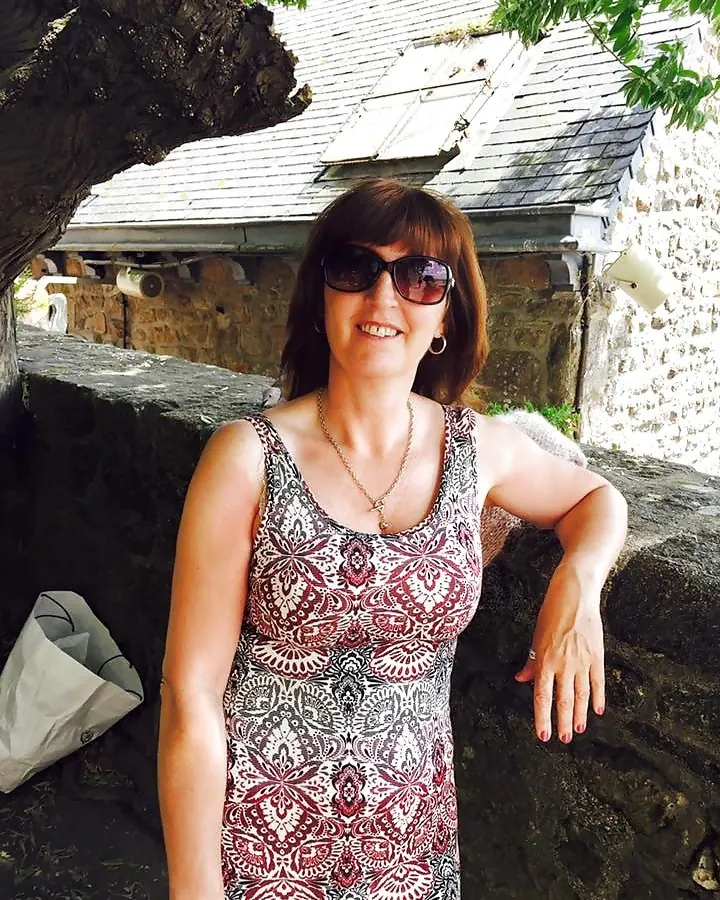 Femme souriante portant des lunettes de soleil et une robe imprimée, adossée à un mur de pierre devant une maison ancienne.