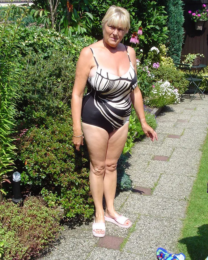 Femme blonde en maillot de bain noir et blanc, debout dans un jardin fleuri, portant des sandales roses.