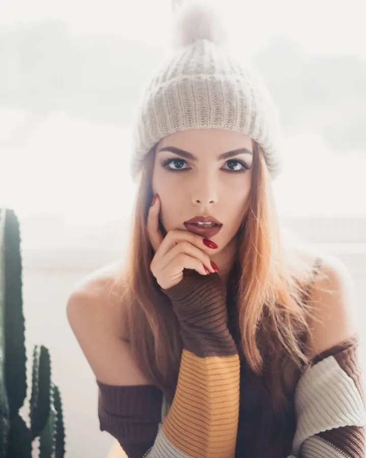 Femme blonde en bonnet tricoté beige, posant près d'un cactus, la main sur le menton, regardant l'objectif avec intensité.
