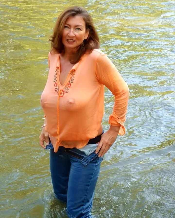 Femme aux cheveux bruns portant un haut orange et un jean, debout dans l'eau.