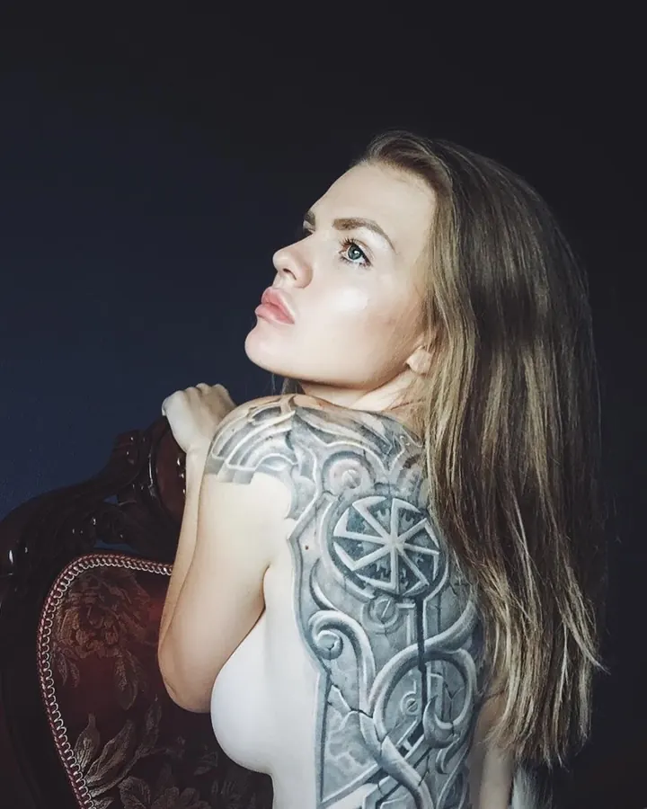 Femme aux cheveux bruns portant un haut blanc et un sous-vêtement noir, avec un tatouage géométrique sur le dos, posant près d'une chaise ancienne.