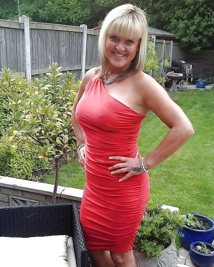 Femme blonde aux yeux bleus, souriante, portant une robe rouge ajustée, debout dans un jardin verdoyant.