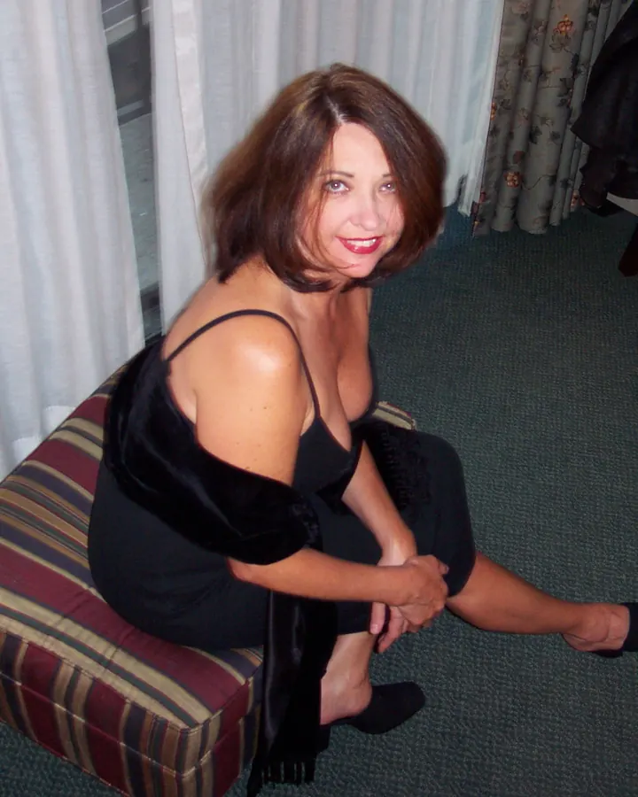 Femme blonde aux cheveux courts, en maillot de bain orange, assise sur un ponton, buvant un cocktail, souriante.