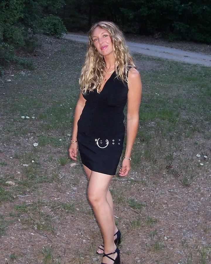 Femme blonde en haut rose à dentelle, posant près d’un lit
