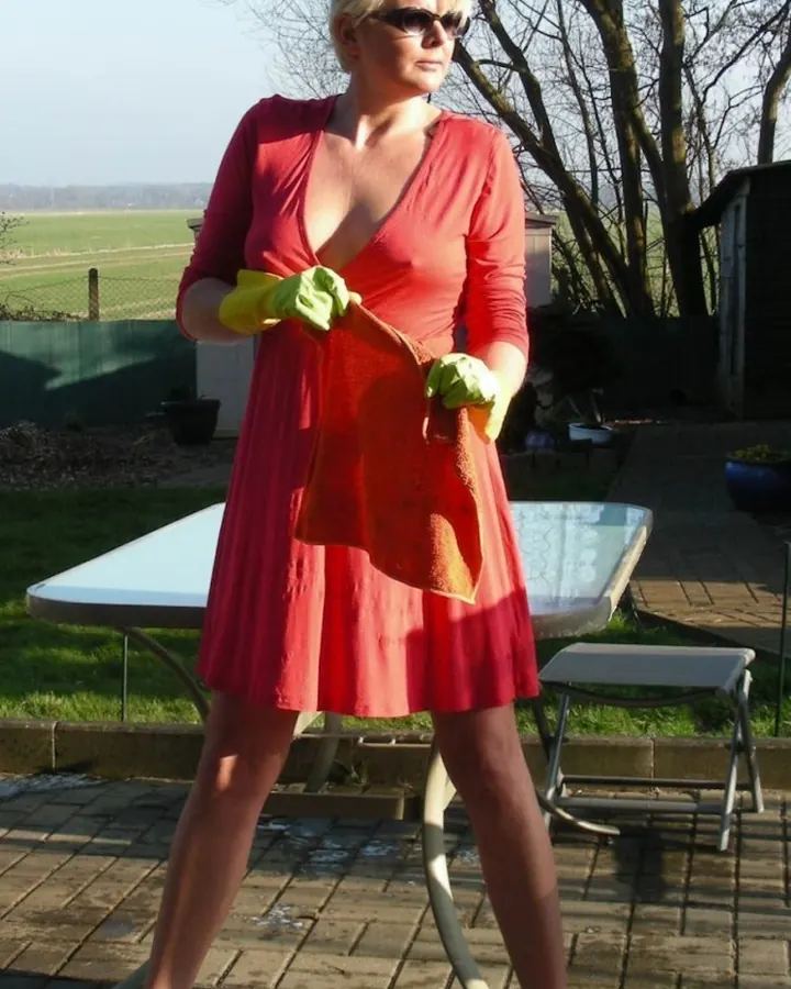 Femme blonde en robe rouge, portant des gants jaunes, nettoyant une table de jardin