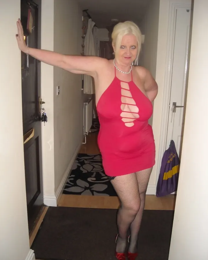 Femme blonde aux yeux bleus portant une robe rouge fendue, debout dans un couloir avec des collants noirs et des chaussures rouges.