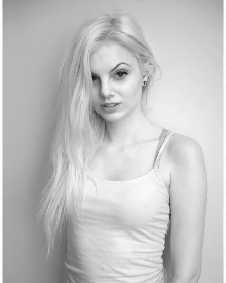 Jeune femme blonde aux cheveux blonds portant un maillot de bain noir, posant de dos près d'une piscine.