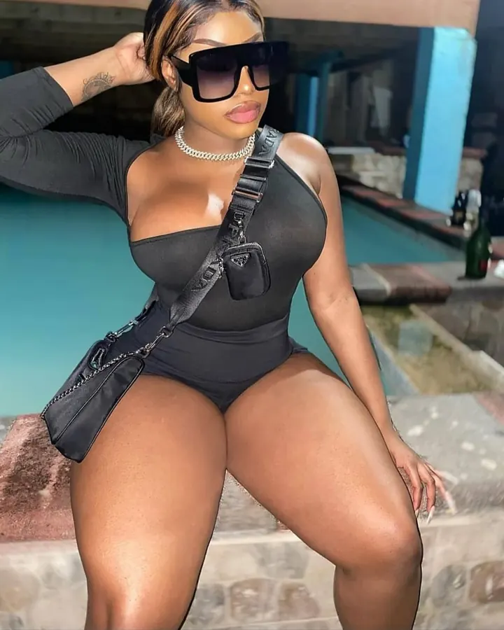 Femme aux cheveux châtains portant un body noir, des lunettes de soleil, un collier en chaîne, posant près d'une piscine, une main dans les cheveux.