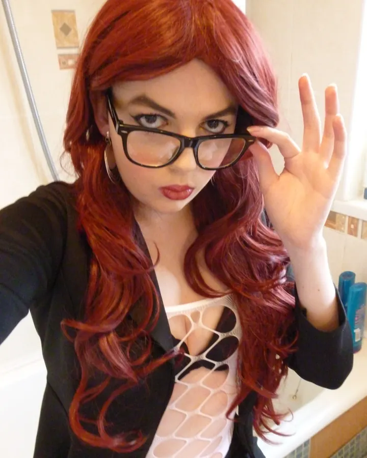 Femme aux cheveux roux en haut noir transparent, blazer noir, lunettes, posant dans une salle de bain