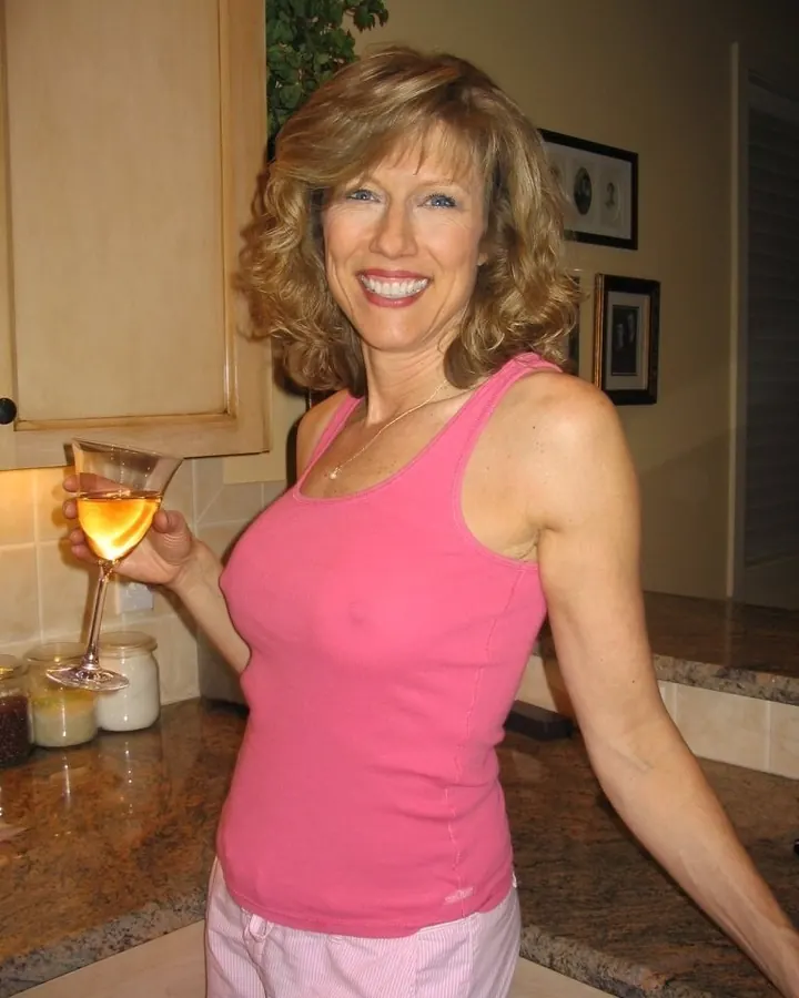 Femme aux cheveux blonds ondulés, souriante, tenant un verre de vin blanc, vêtue d'un haut rose, debout dans une cuisine.