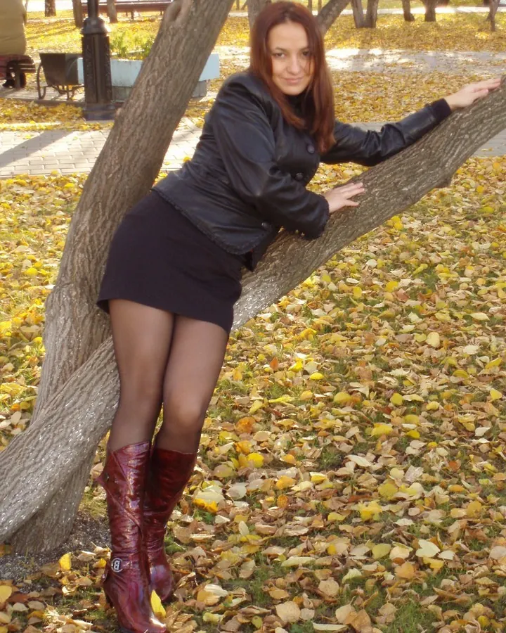 Femme aux cheveux roux mi-longs, souriante, vêtue d'un haut noir, debout dans une pièce avec un sol en bois.