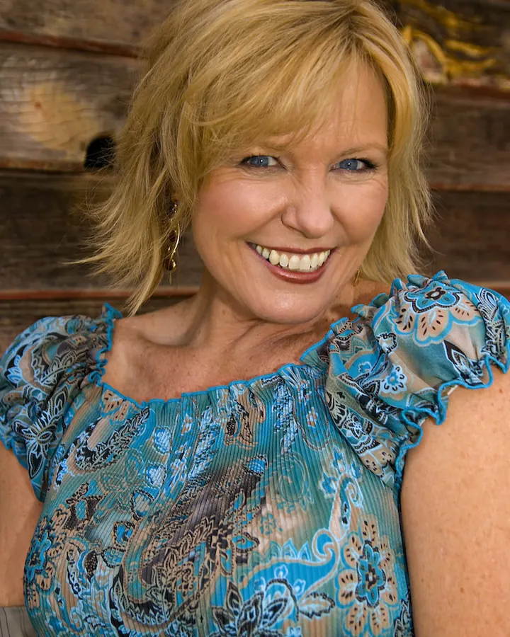 Femme aux cheveux blonds courts, souriante, vêtue d'une robe bleue à motifs, debout devant un mur en bois.
