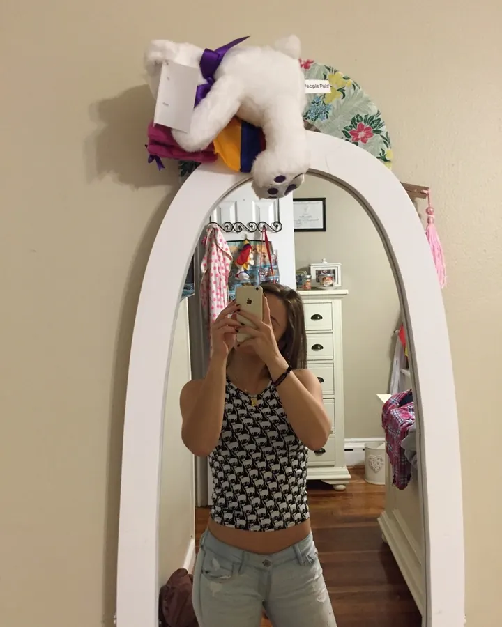 Jeune femme prenant un selfie dans un miroir blanc, portant un haut à motifs animaux et un jean déchiré, avec un ours en peluche sur le miroir.