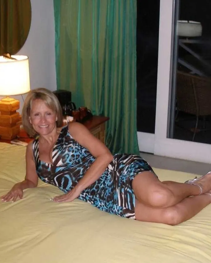 Femme blonde souriante en robe imprimée, allongée sur un lit jaune avec une lampe à côté.
