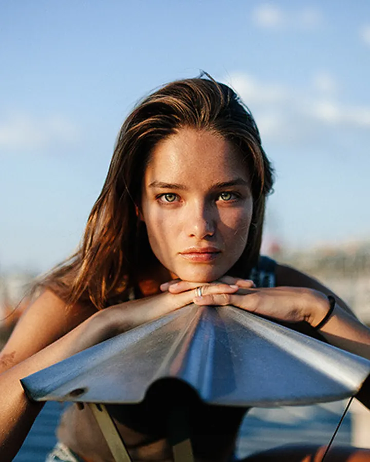 Jeune femme brune en maillot de bain noir, regardant vers l'horizon depuis un balcon avec vue sur la mer et une plage.
