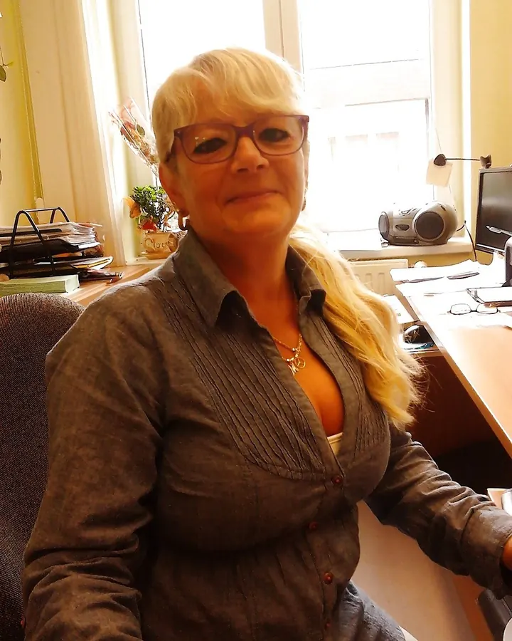 Femme blonde assise à un bureau, portant une chemise grise boutonnée, avec des cheveux longs et des lunettes.