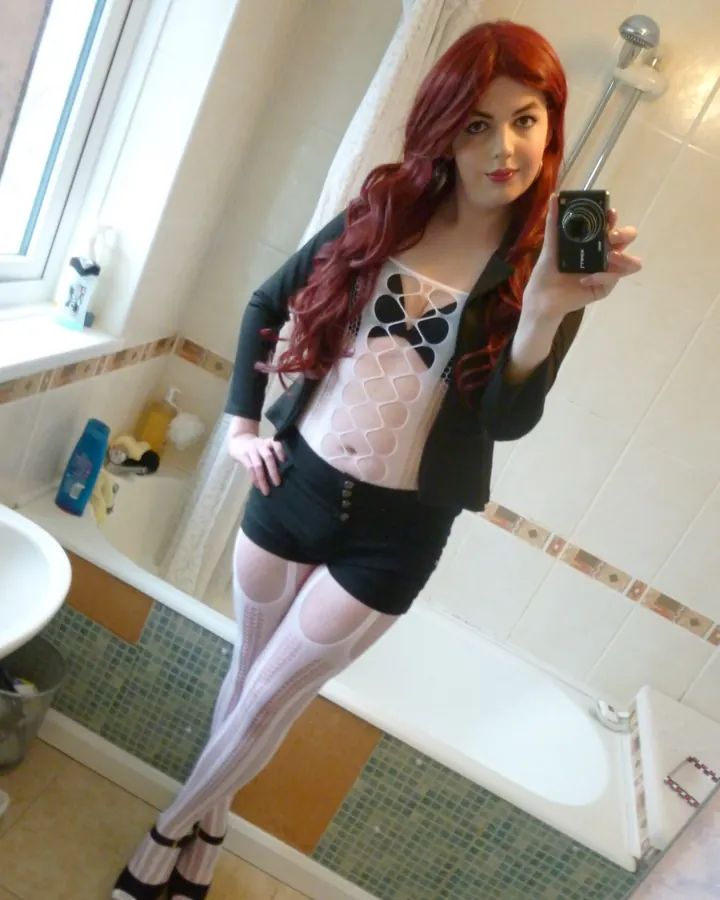 Femme aux cheveux roux, portant un haut noir et un short noir, posant dans une salle de bain.