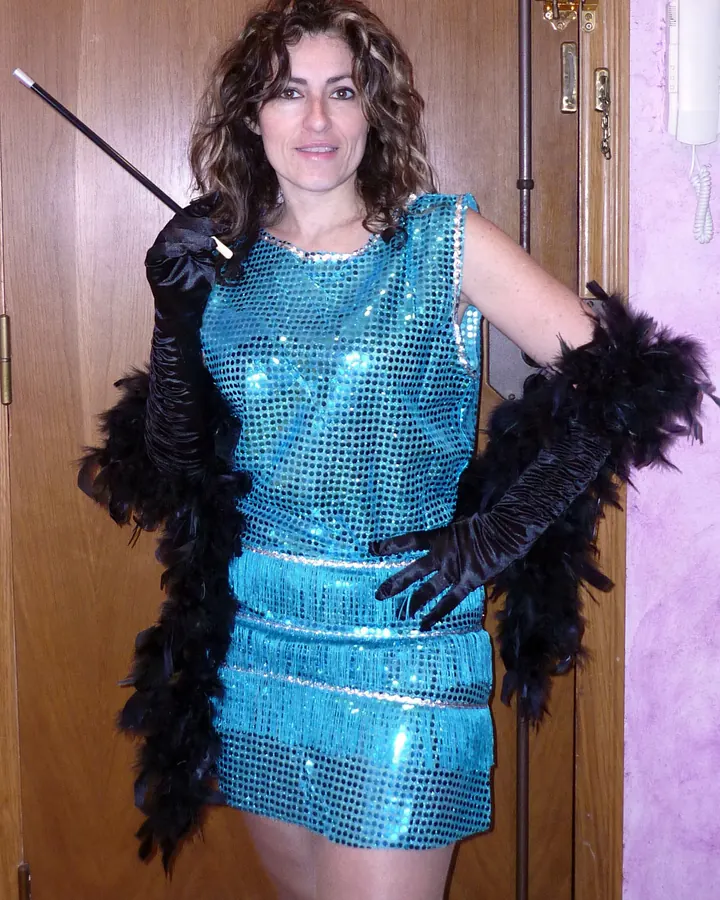 Femme en robe bleue pailletée, gants noirs, tenant un fouet, devant une porte.