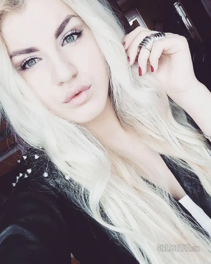 Jeune femme aux cheveux blonds, portant un haut noir et un short noir, assise sur un canapé, souriant à la caméra.