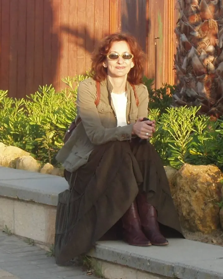Femme aux cheveux châtains assise sur un banc, portant une veste beige, une jupe longue foncée et des bottes marron, devant un mur en bois et des buissons verts.