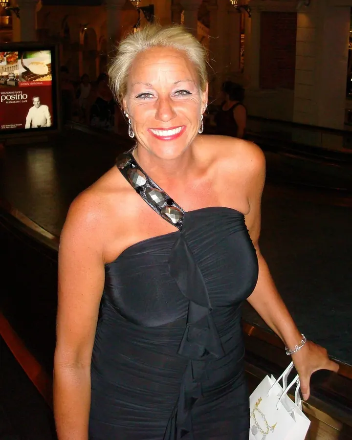 Femme blonde en robe noire asymétrique, souriante, tenant un sac à main dans un hall élégant.