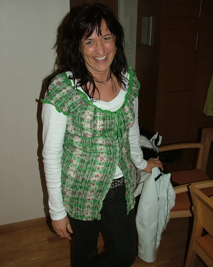 Femme aux cheveux bruns-noirs courts, yeux bleus, portant un haut à col en V, souriant légèrement, debout en extérieur.