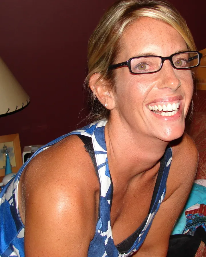 Femme aux cheveux blonds mi-longs, yeux gris, portant un haut à col en V, souriant légèrement, debout en intérieur.