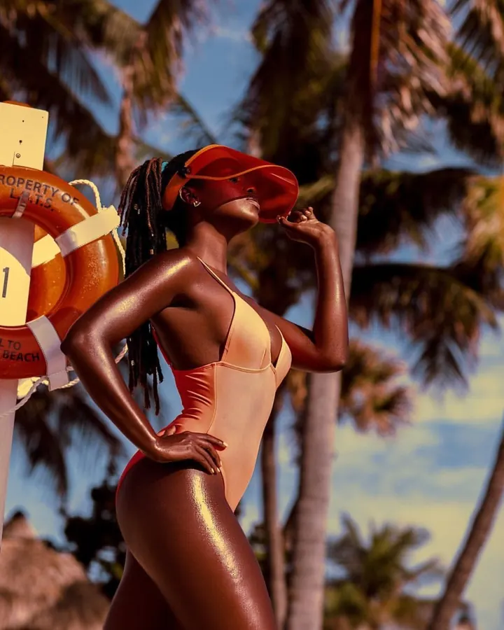 Jeune femme africaine aux cheveux bruns-noirs longs, portant un maillot de bain orange, tenant un casque de protection rouge, debout sur une plage avec des palmiers.