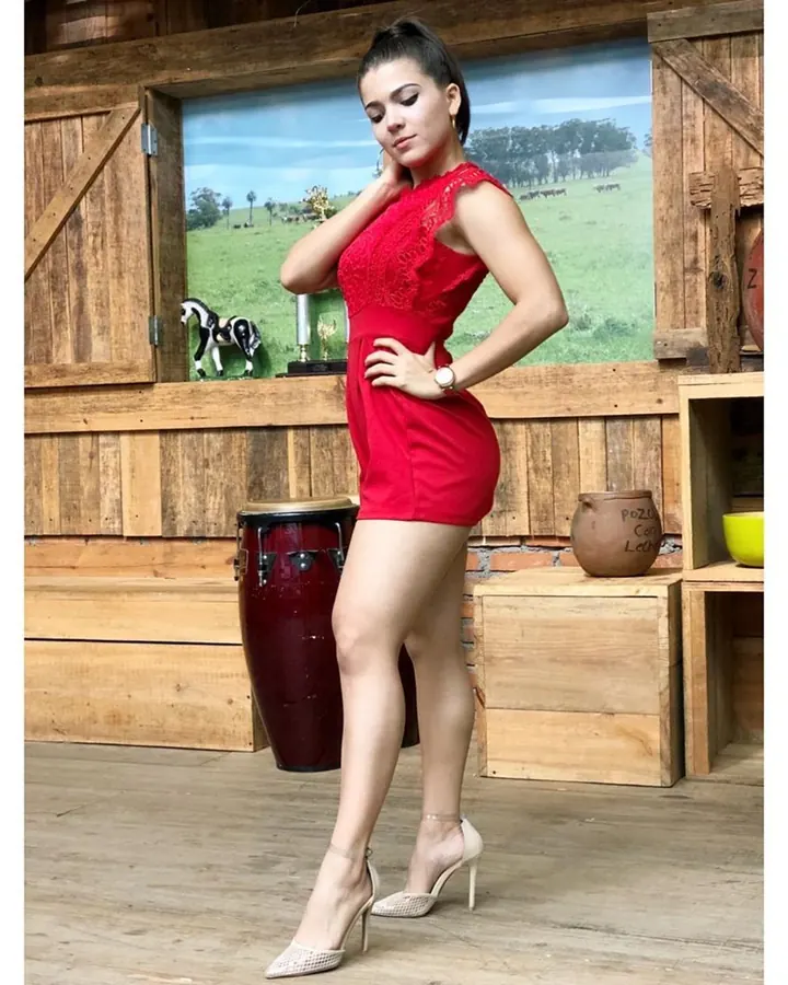 Femme blonde portant une veste en cuir noir, un short en cuir, des collants noirs et des bottes hautes, posant près d'une porte blanche.