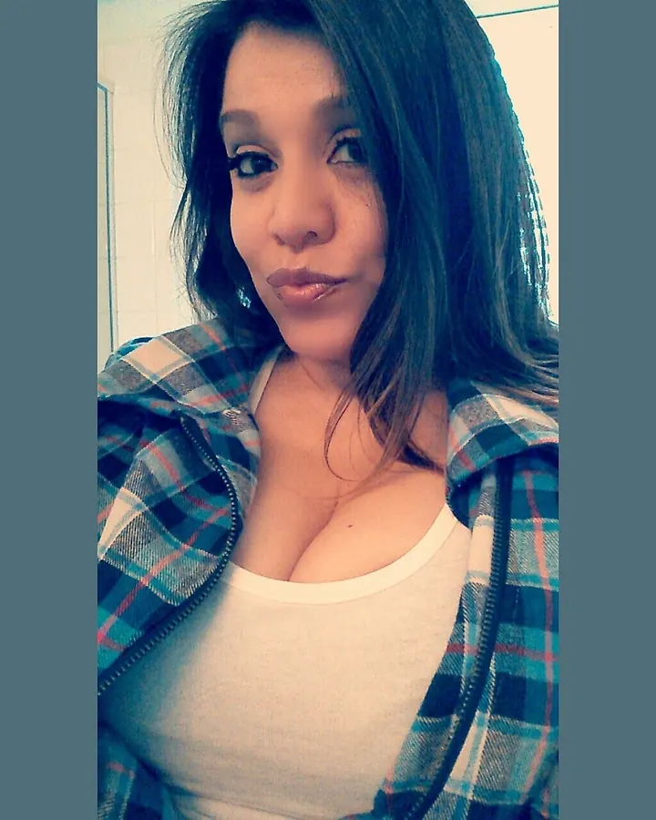 Femme blonde portant une robe rouge fendue, tenant un téléphone doré, prenant un selfie devant un mur en bois foncé.