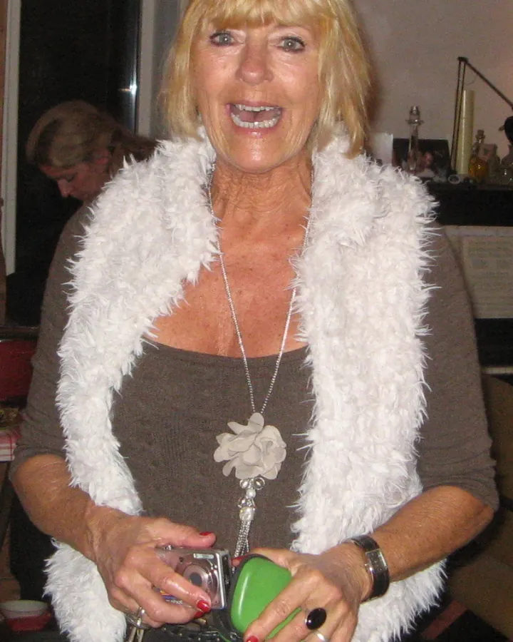 Femme blonde plus âgée souriante, portant une veste en fourrure blanche, tenant un appareil photo numérique et un petit sac vert.