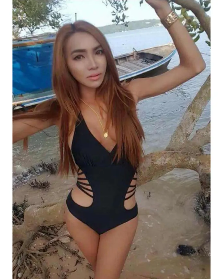 Femme aux cheveux bruns-noirs longs, portant un maillot de bain noir avec découpes latérales, debout près d'une rivière avec un bateau en arrière-plan.