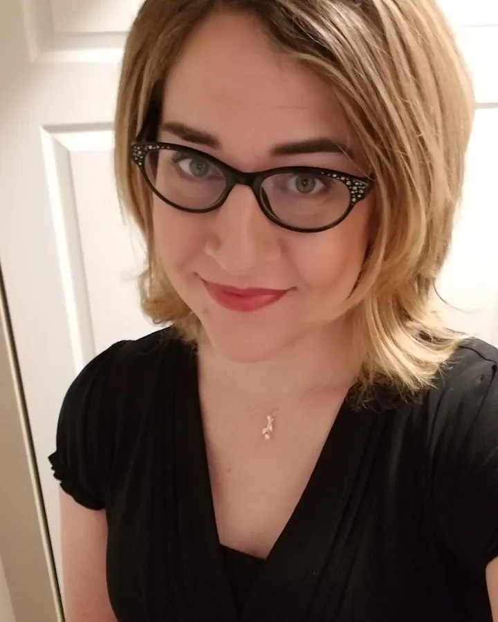 Femme aux cheveux blonds, en haut noir et short gris, prenant un selfie devant un miroir dans une chambre.