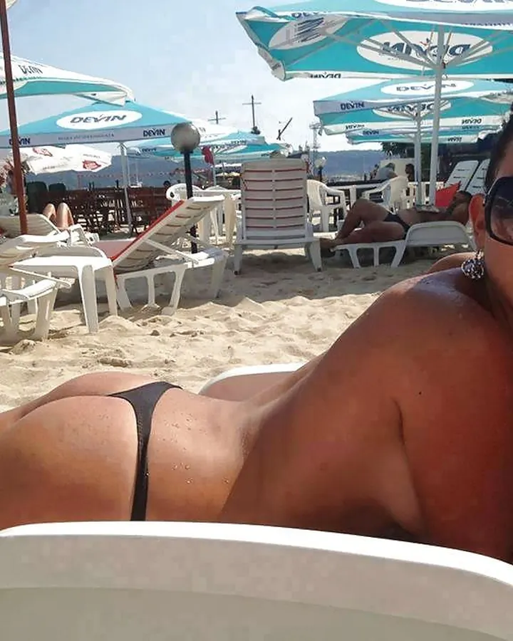 Femme brune en maillot noir sur une plage ensoleillée, sous un parasol bleu, regardant l'appareil.