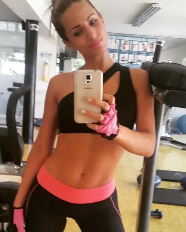 Femme en tenue de sport noire et rose, prenant un selfie dans une salle de gym.