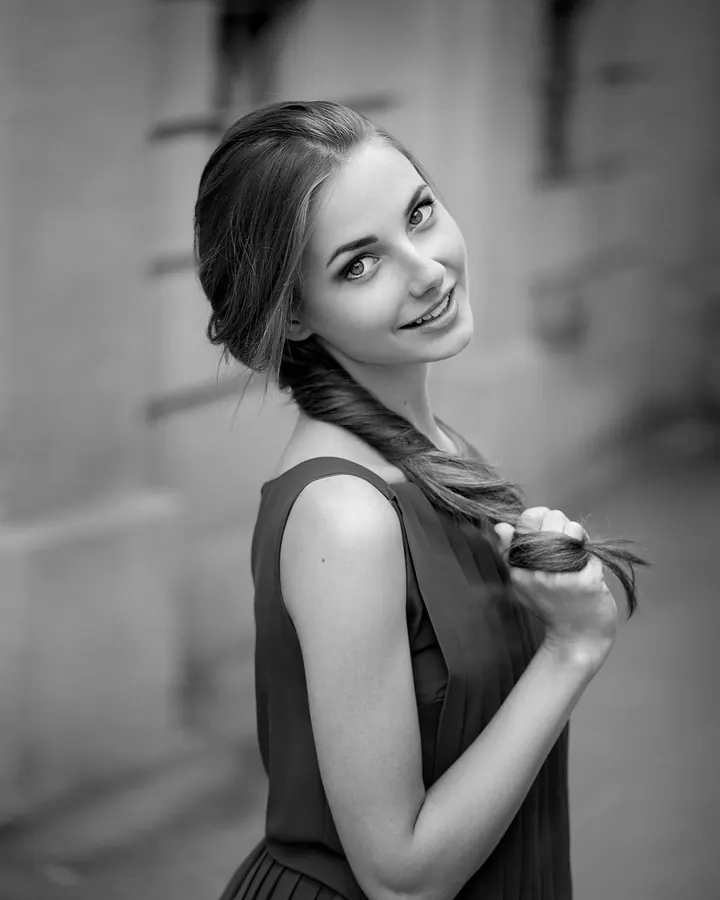 Jeune femme blonde aux yeux bleus, souriante, vêtue d'un haut blanc, posant dans une pièce sombre.