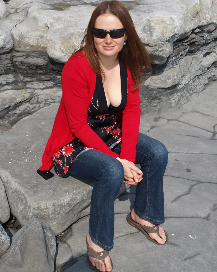 Jeune femme assise sur des rochers, vêtue d’un cardigan rouge, d’une robe fleurie et de jeans, souriant à l’appareil.
