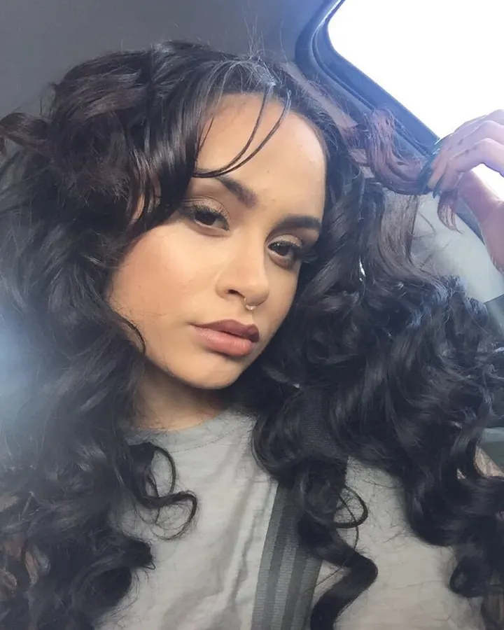 Femme aux cheveux bruns, en haut noir à strass, jeans, prenant un selfie dans une pièce.