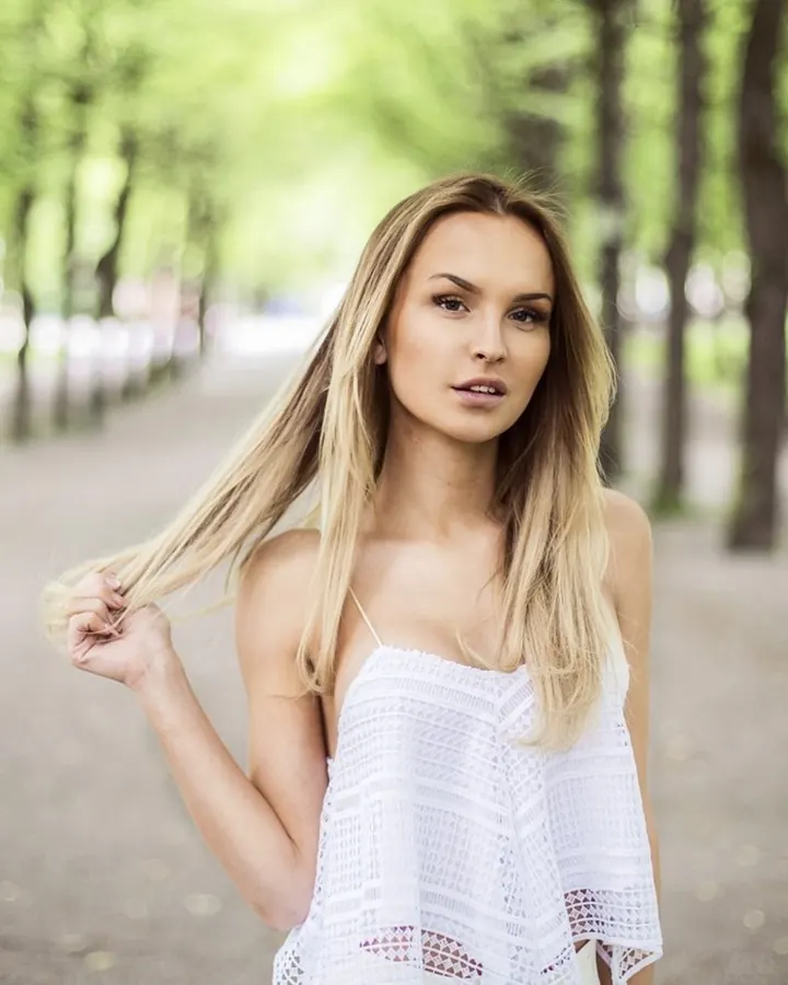 Femme blonde portant une robe blanche, marchant dans un parc arboré, tenant une mèche de cheveux.
