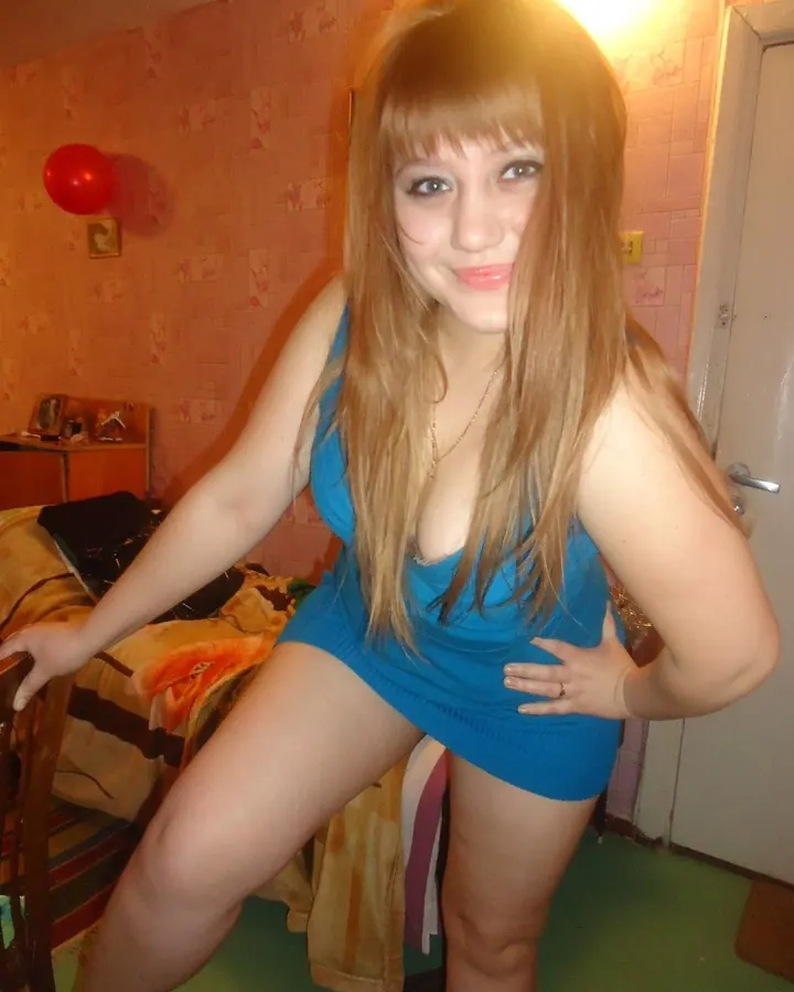 Femme aux cheveux bruns portant une robe bleue, posant dans une chambre avec un ballon rouge en arrière-plan.
