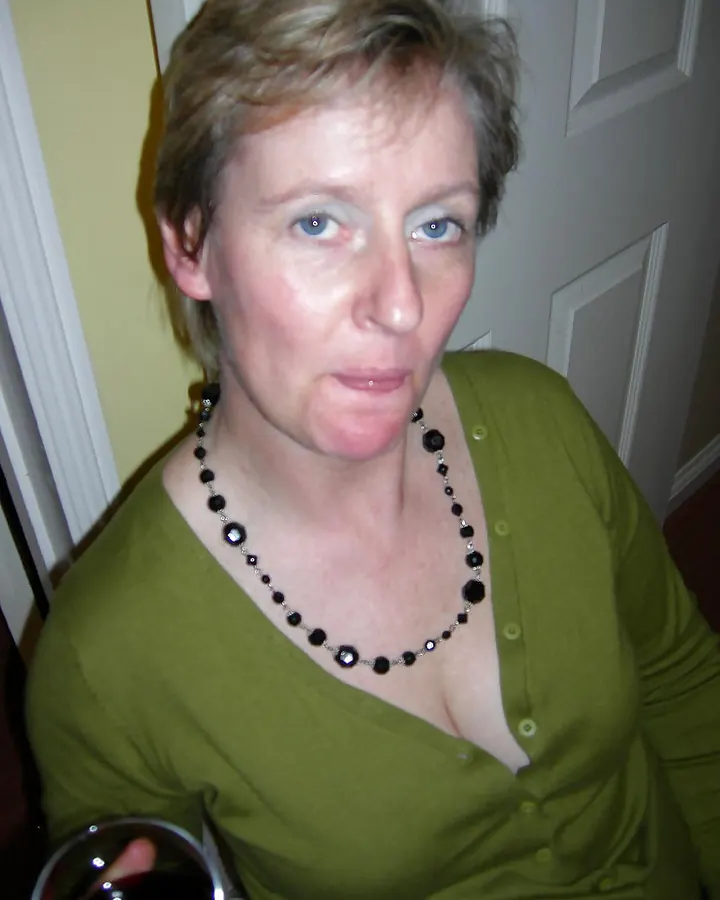 Femme aux cheveux courts blonds portant un haut vert, regardant l'appareil photo dans une pièce intérieure.