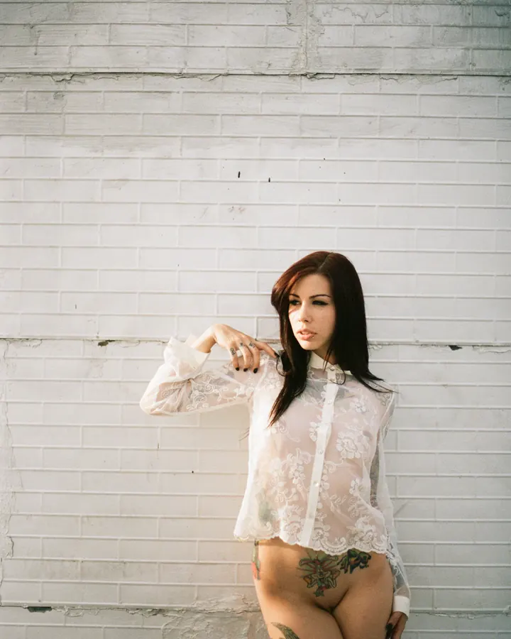 Femme aux cheveux bruns portant une chemise blanche transparente, posant contre un mur blanc, tatouages visibles