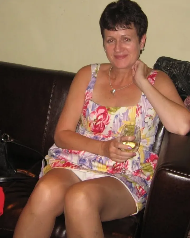 Femme aux cheveux bruns, portant une robe noire, assise sur un canapé en cuir brun, regardant directement l'appareil photo.