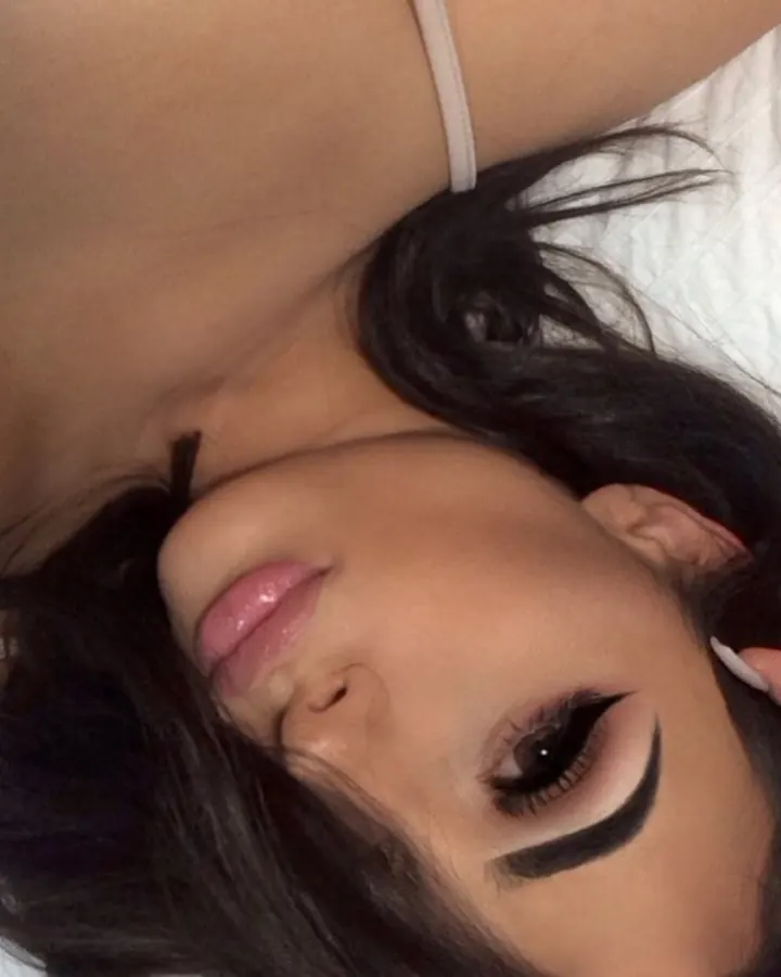 Femme aux cheveux noirs, maquillée avec un trait d'eye-liner épais, allongée sur un lit blanc, main dans les cheveux.
