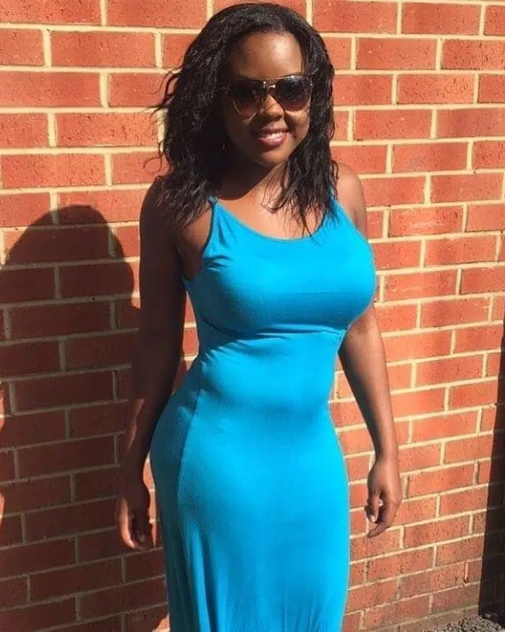Femme noire en robe bleue ajustée, posant devant un mur de briques rouges, portant des lunettes de soleil