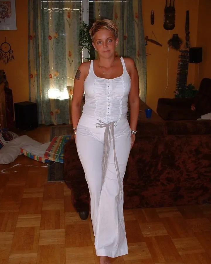Femme aux cheveux blonds, en haut blanc et pantalon noir, allongée sur un lit, souriant, main sur la joue
