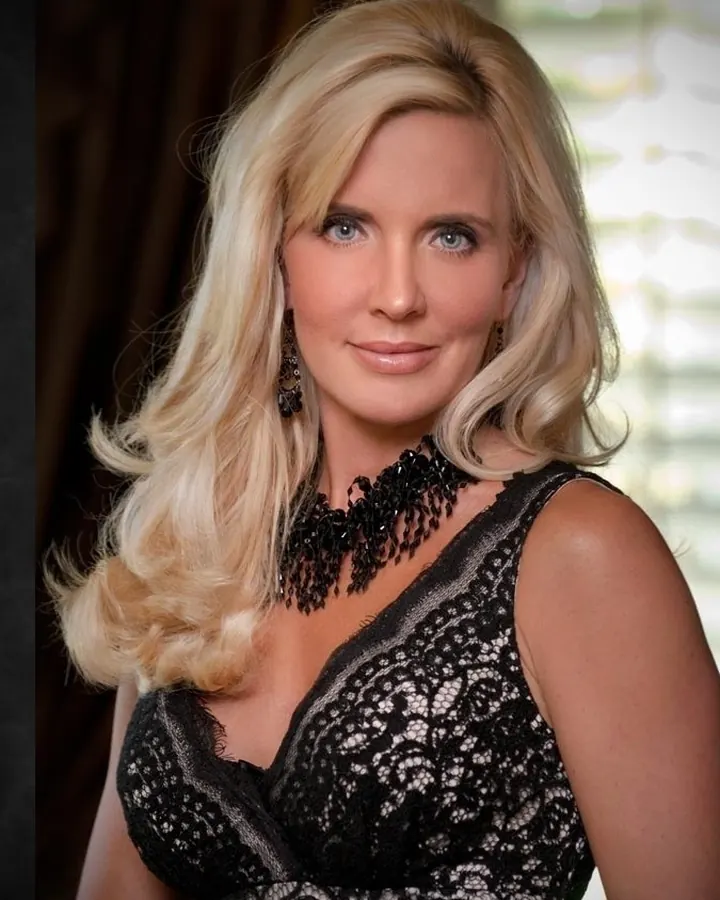 Femme blonde aux yeux bleus portant une robe noire en dentelle avec un collier noir élaboré, souriant légèrement à l'appareil.